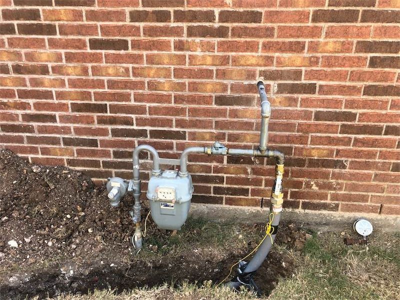 Gas meter and pipes against a brick wall, in a grassy area with exposed soil.