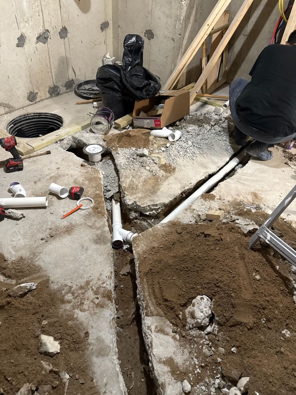 Plumbing work in a basement. A person works on white pipes in a concrete trench with dirt.