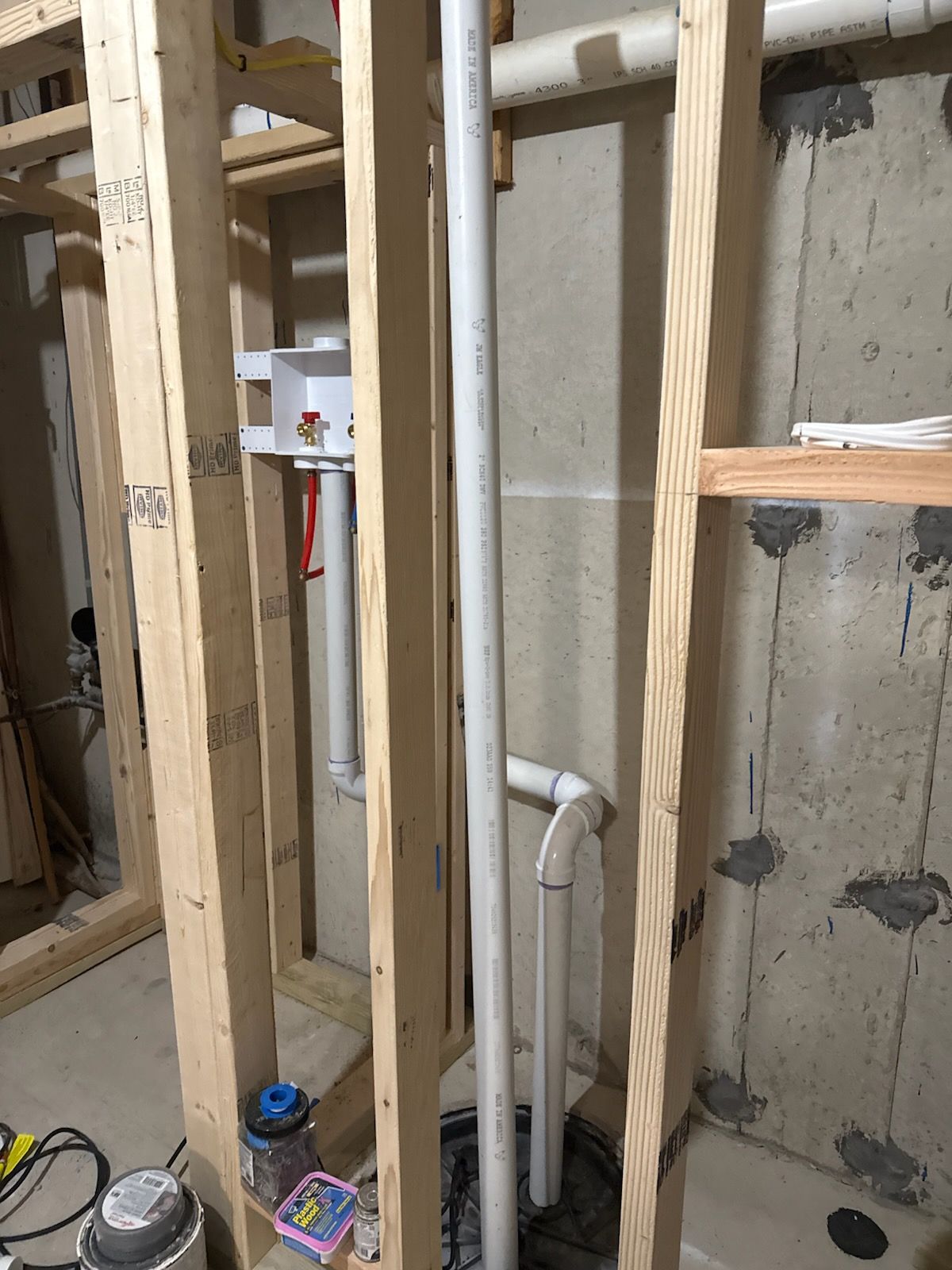 Framing with plumbing visible. White pipes run down concrete wall; electrical box on left.