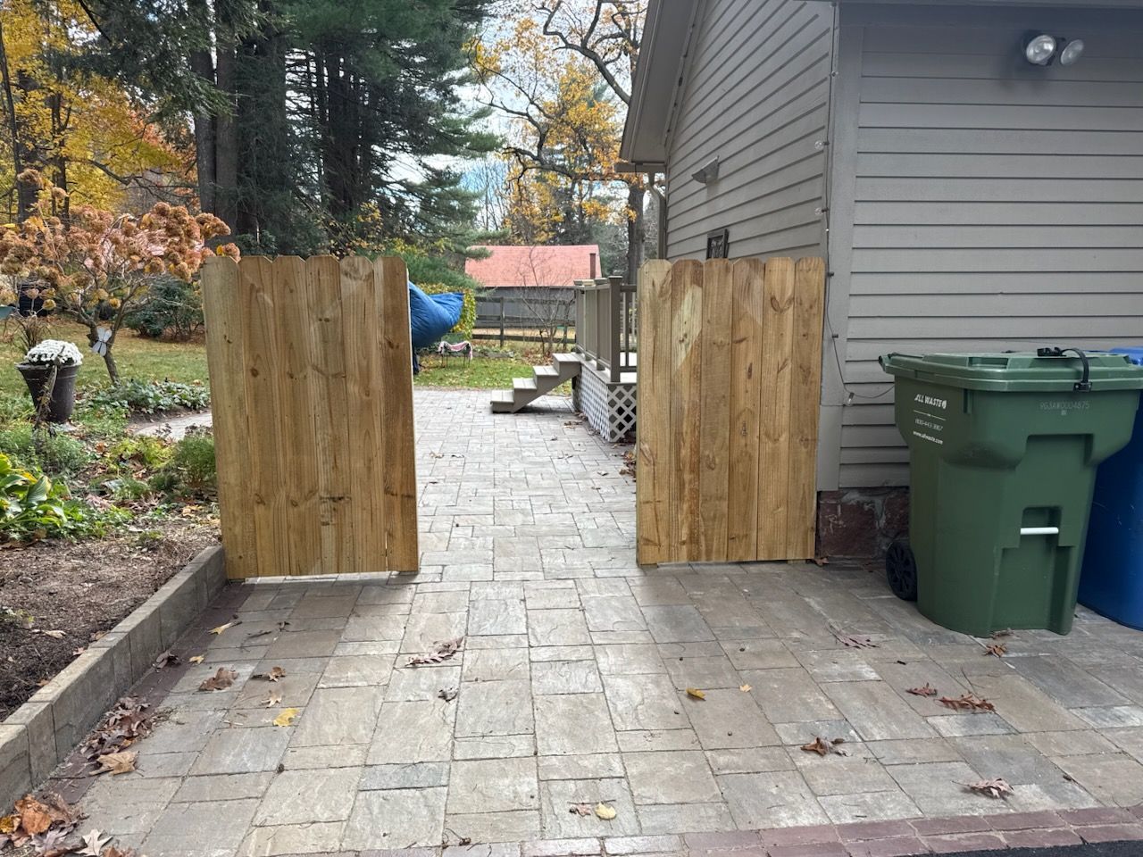 Two wooden gates on a paved patio lead to a backyard. Trash cans stand beside a building.