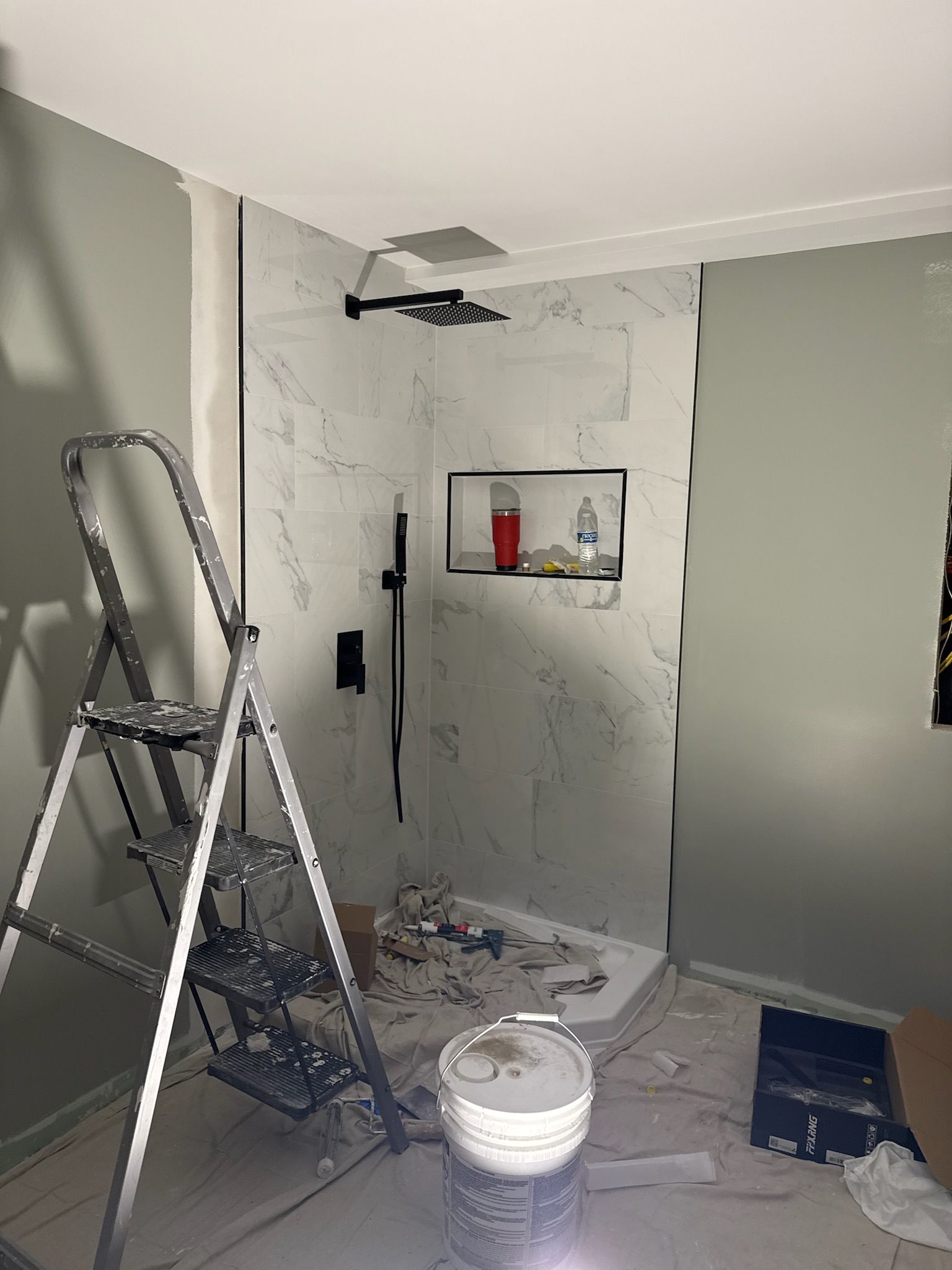 A step ladder next to a partially tiled shower with black fixtures and a built-in shelf; renovation in progress.