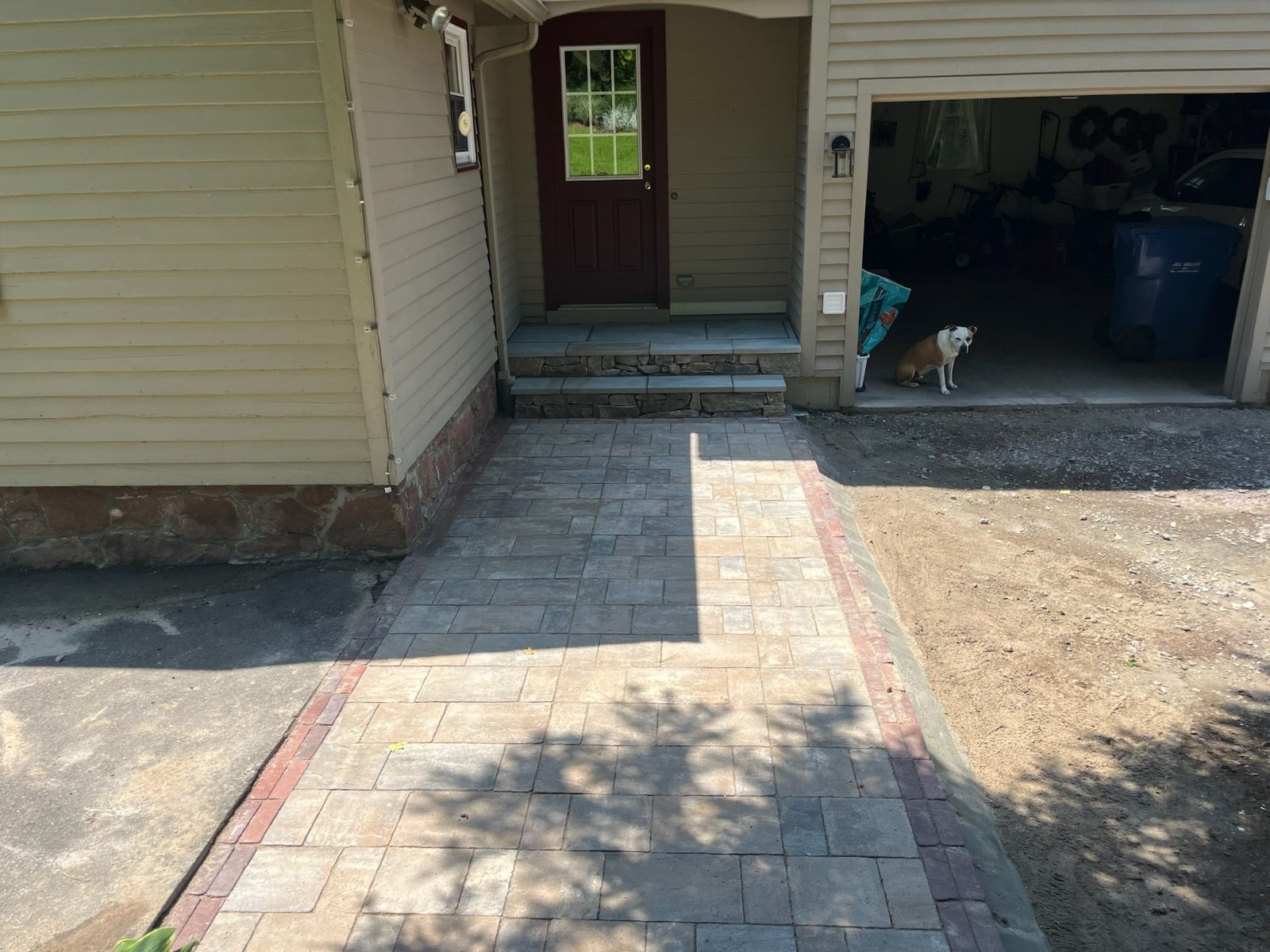Stone walkway leads to house with steps, red door. Open garage with dog.