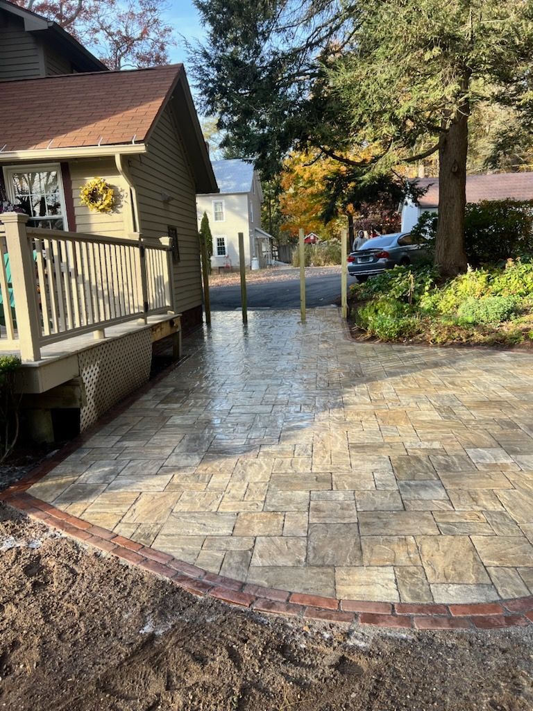 Brick paver patio with a brick border leading to a house, flanked by a wooden porch and a tree.