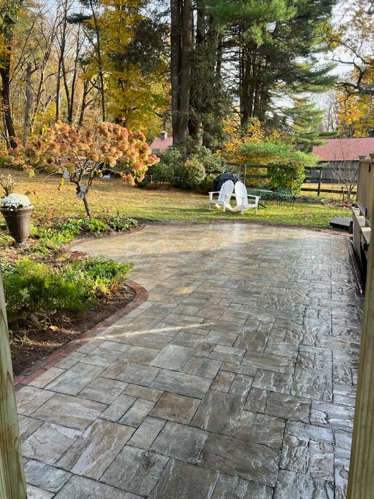 Patio with stone pavers, greenery, and trees in a yard. Two white chairs sit in the background.