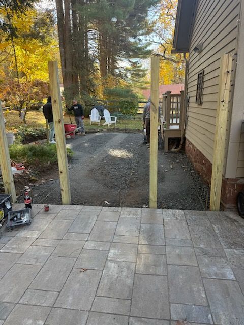 Wooden fence posts being installed next to a paved patio near a house and a gravel driveway.