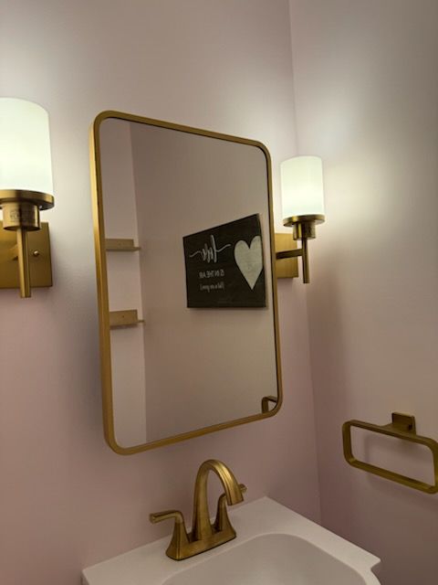 Gold-framed mirror over a sink in a bathroom with gold fixtures and sconces, light pink walls.