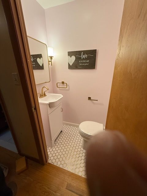 Small pink bathroom with white fixtures, gold hardware, and hexagon tile flooring. A wooden sign says 