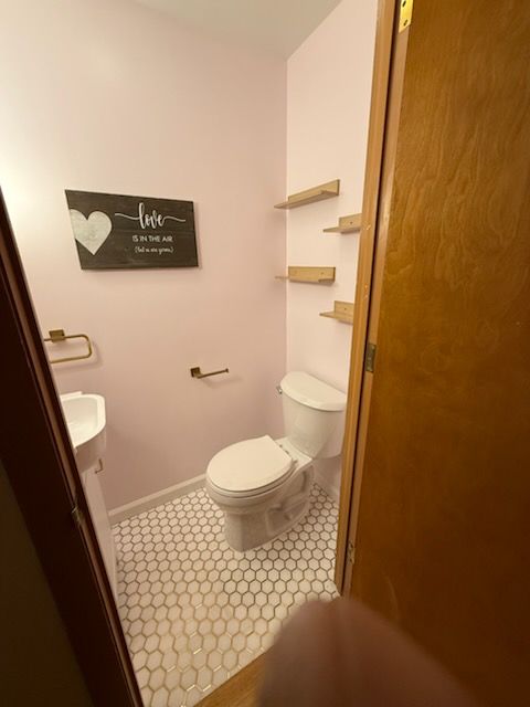 A small, pink-painted bathroom with white tile floor, toilet, and wooden shelves. A 