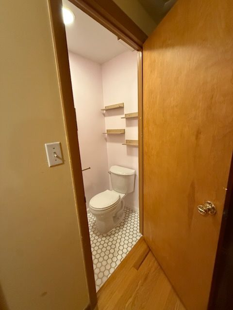Small bathroom with toilet and shelves, viewed from doorway. Pink walls, hexagon tile floor, and wood door.