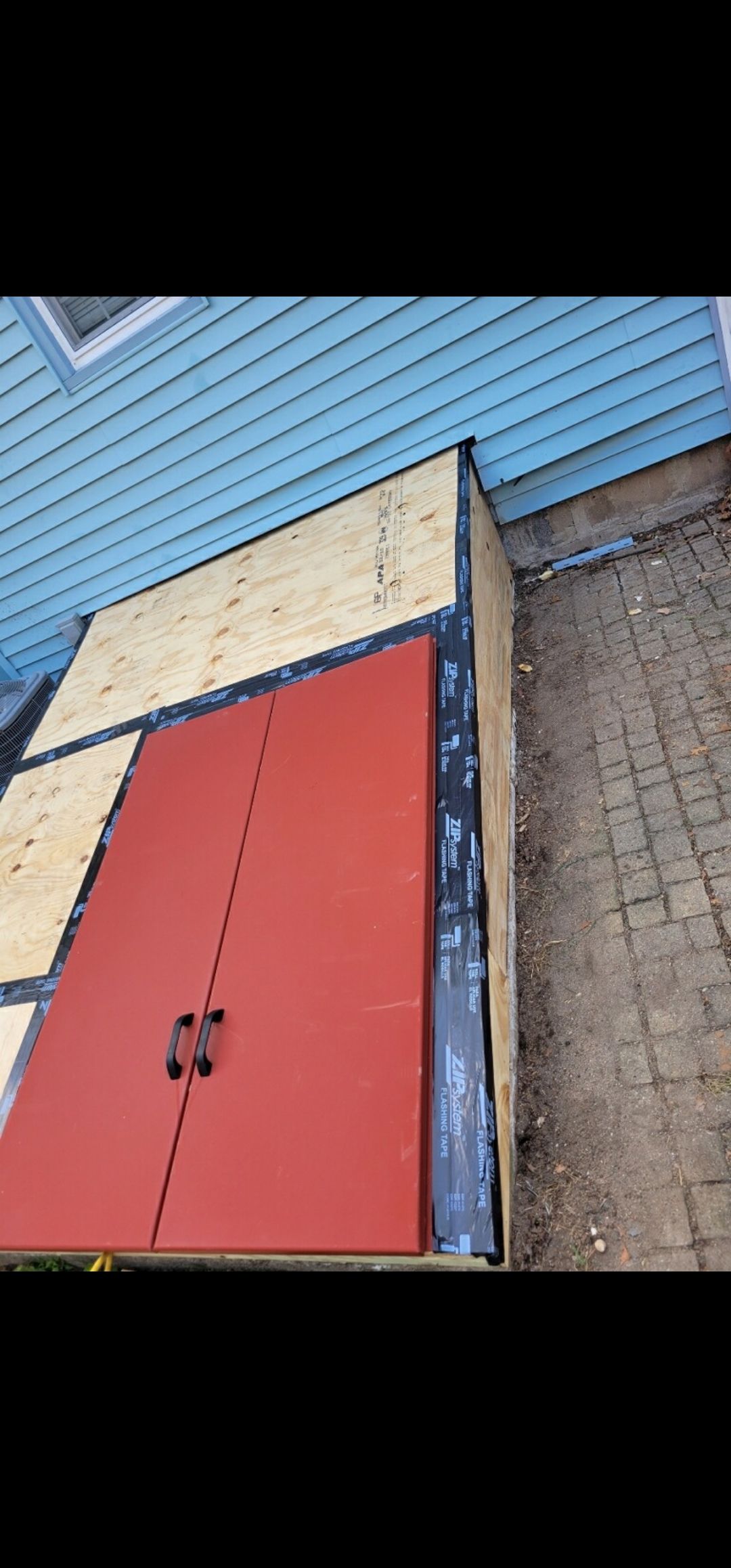 Construction of a red door, possibly for a shed, next to a blue house and concrete patio.