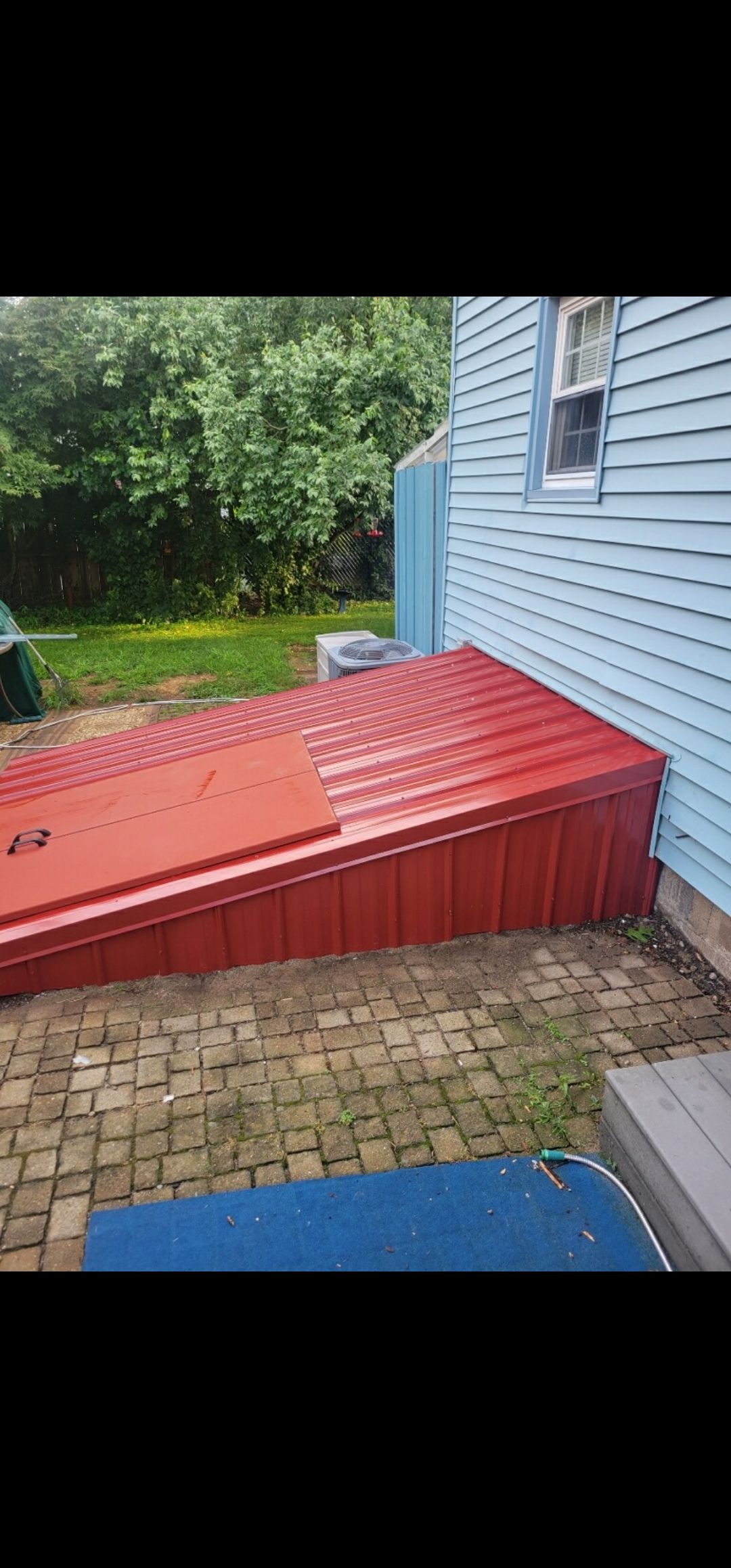 Red entry to a basement in a brick paved area, next to a blue house with a window and a blue entry.