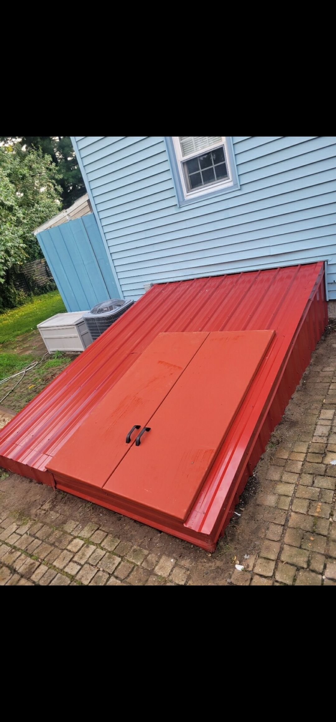 Red metal sheets, likely for a roof, lie on the ground next to a blue house. One sheet has a doorway cutout.