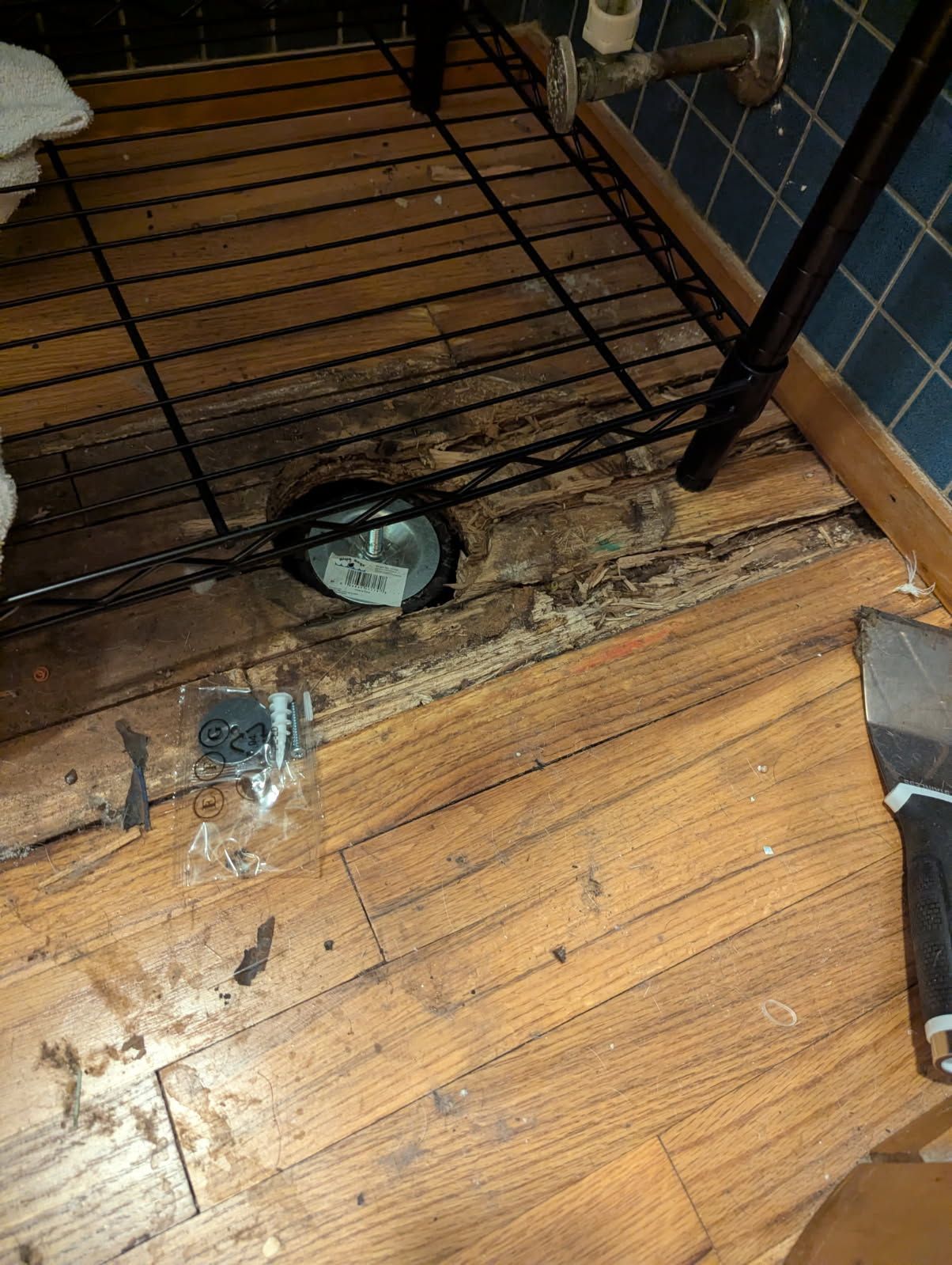 Damaged wooden bathroom floor around a drainpipe, with a wire shelf above it.