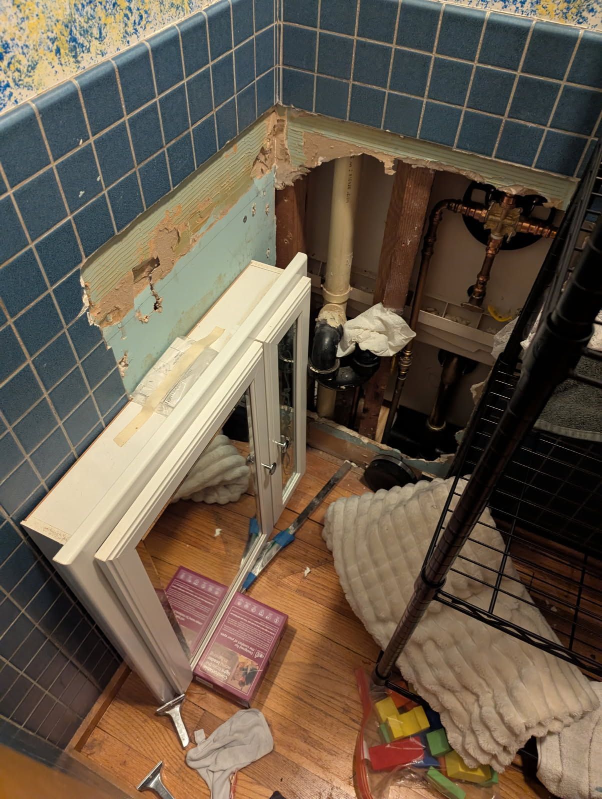 Bathroom wall torn open, revealing pipes and wooden frame; a cabinet door lies on the floor.