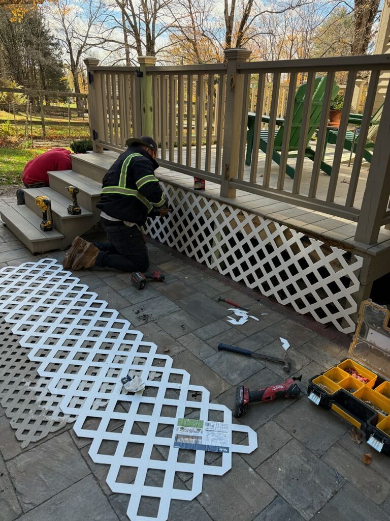 Person installing white lattice on deck. Tools, pavers, and another person visible.