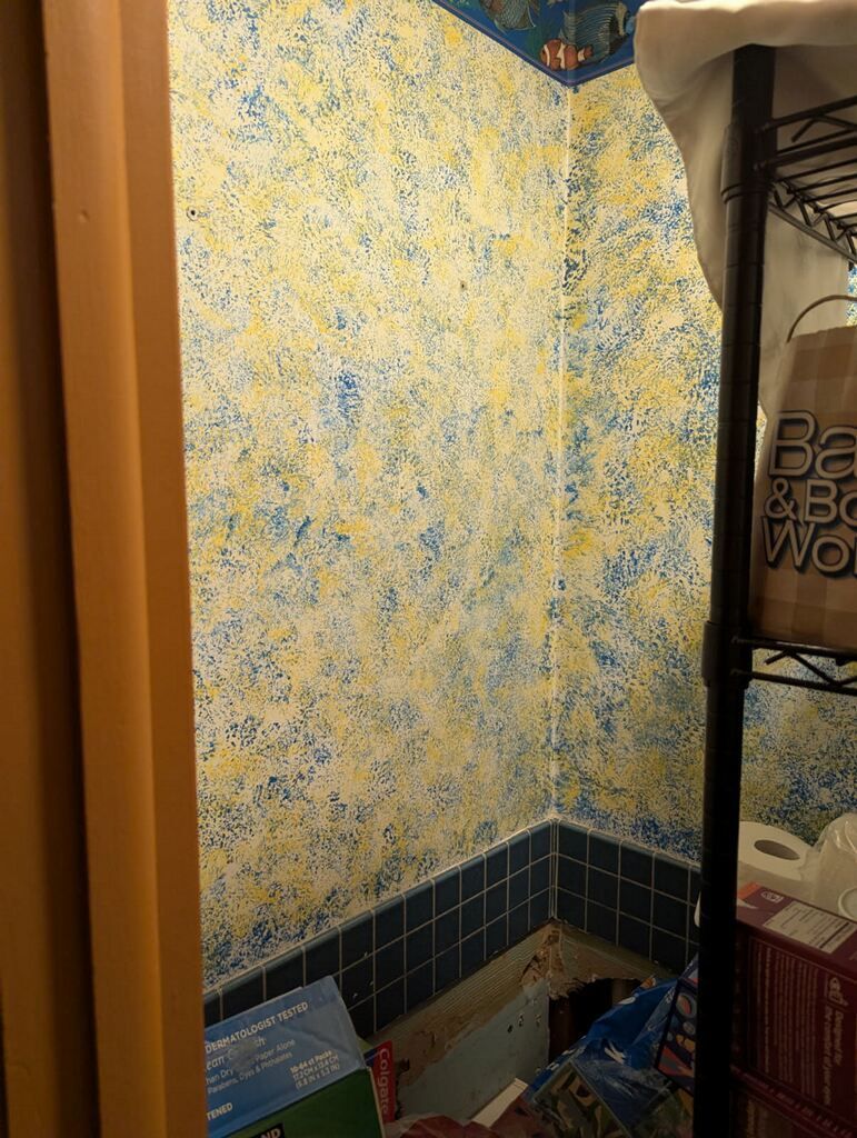 A cluttered small room with textured yellow and blue walls, black shelving, and blue-tiled base.