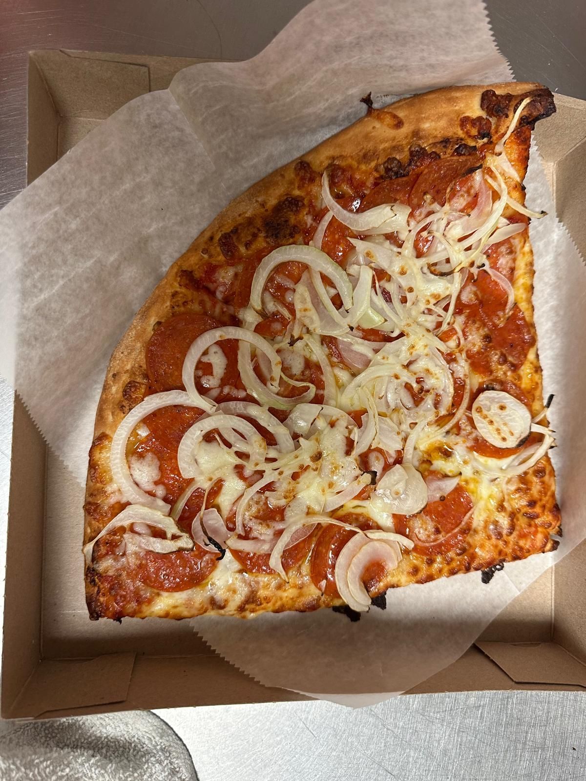 Slice of pizza with pepperoni, onions, and cheese in a cardboard box.