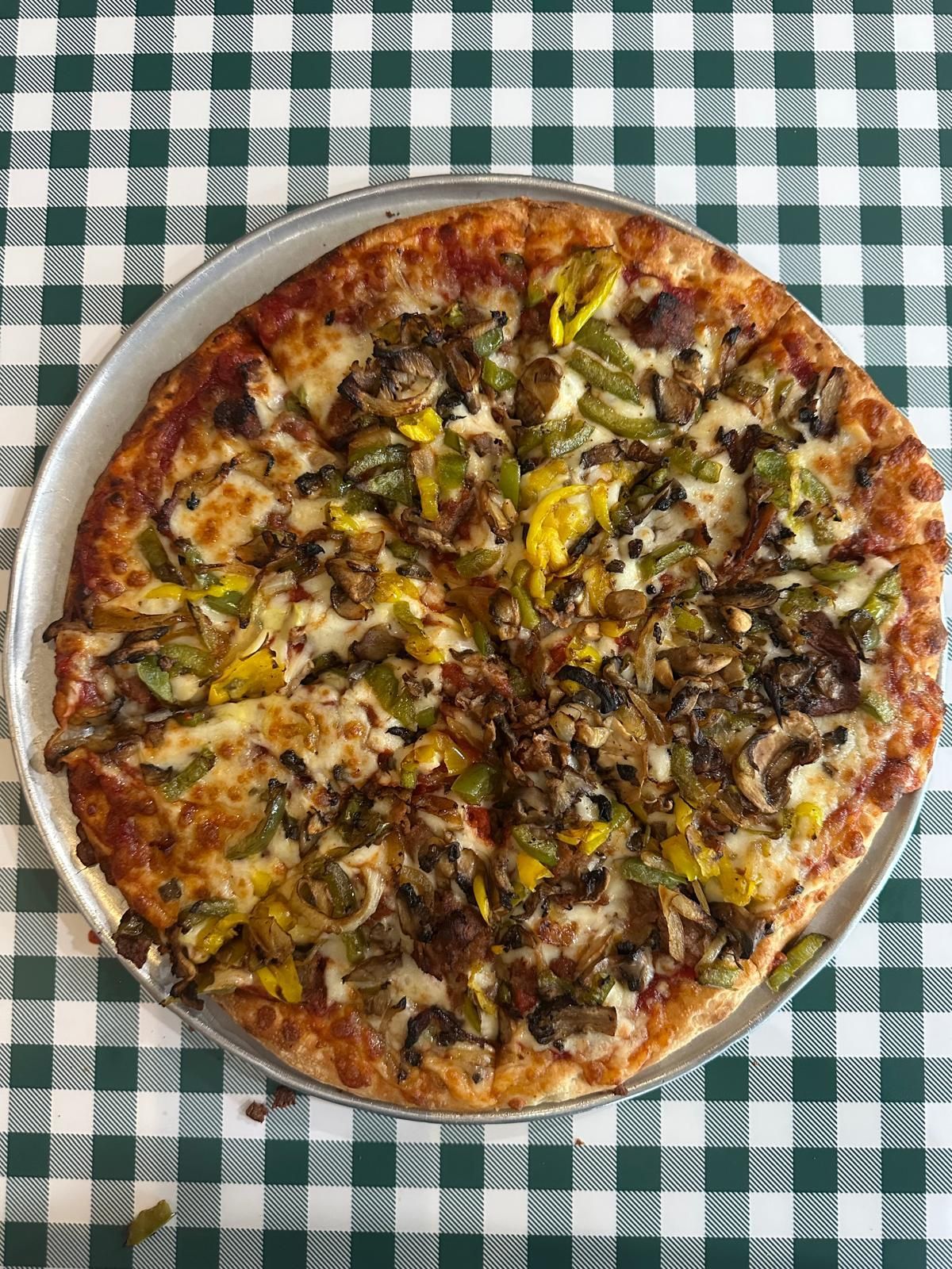 Pizza on a metal tray, topped with vegetables, on a green and white checkered tablecloth.
