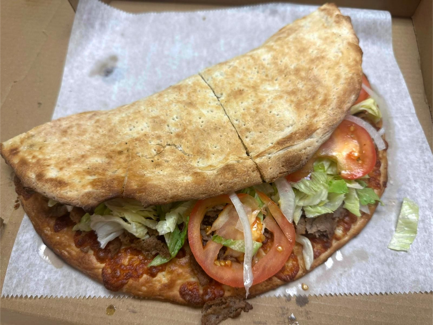 Pizza-like sandwich with lettuce, tomato, and onion in an open cardboard box.