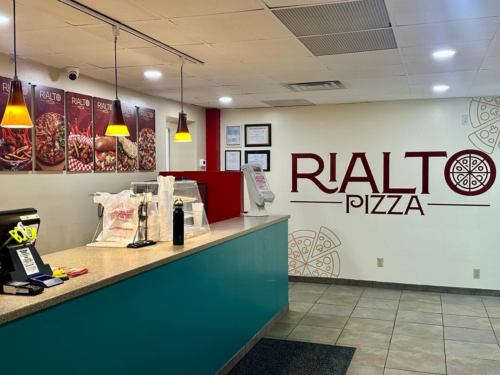 Interior of Rialto Pizza restaurant. Counter with teal front, menu posters, and logo on wall.