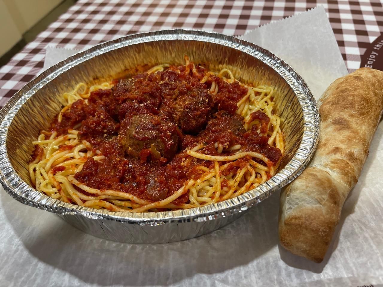 Spaghetti and meatballs with red sauce in a foil container, with bread on a checkered table.