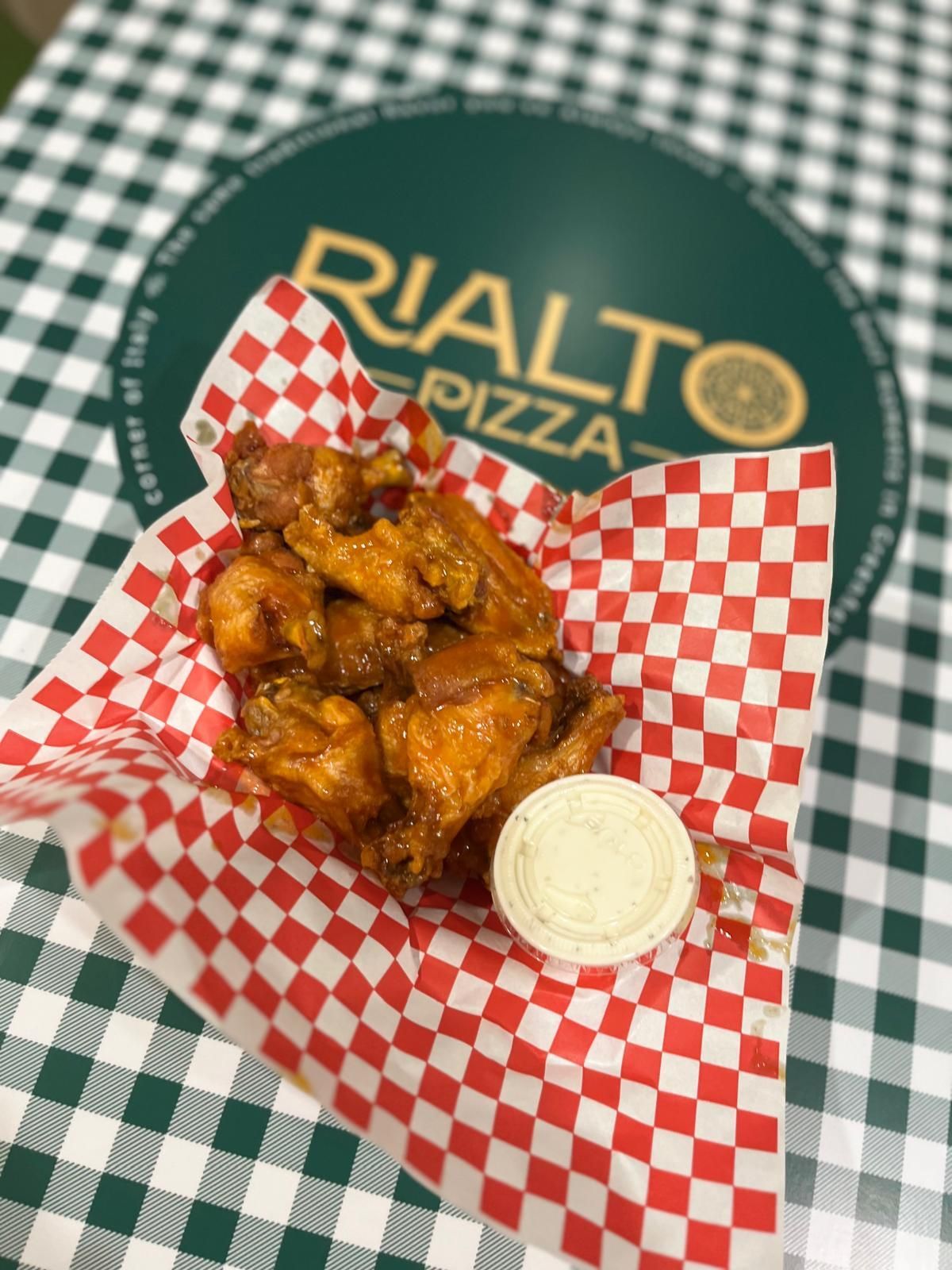 Chicken wings in a checkered basket with dipping sauce, in front of a Rialto Pizza sign.