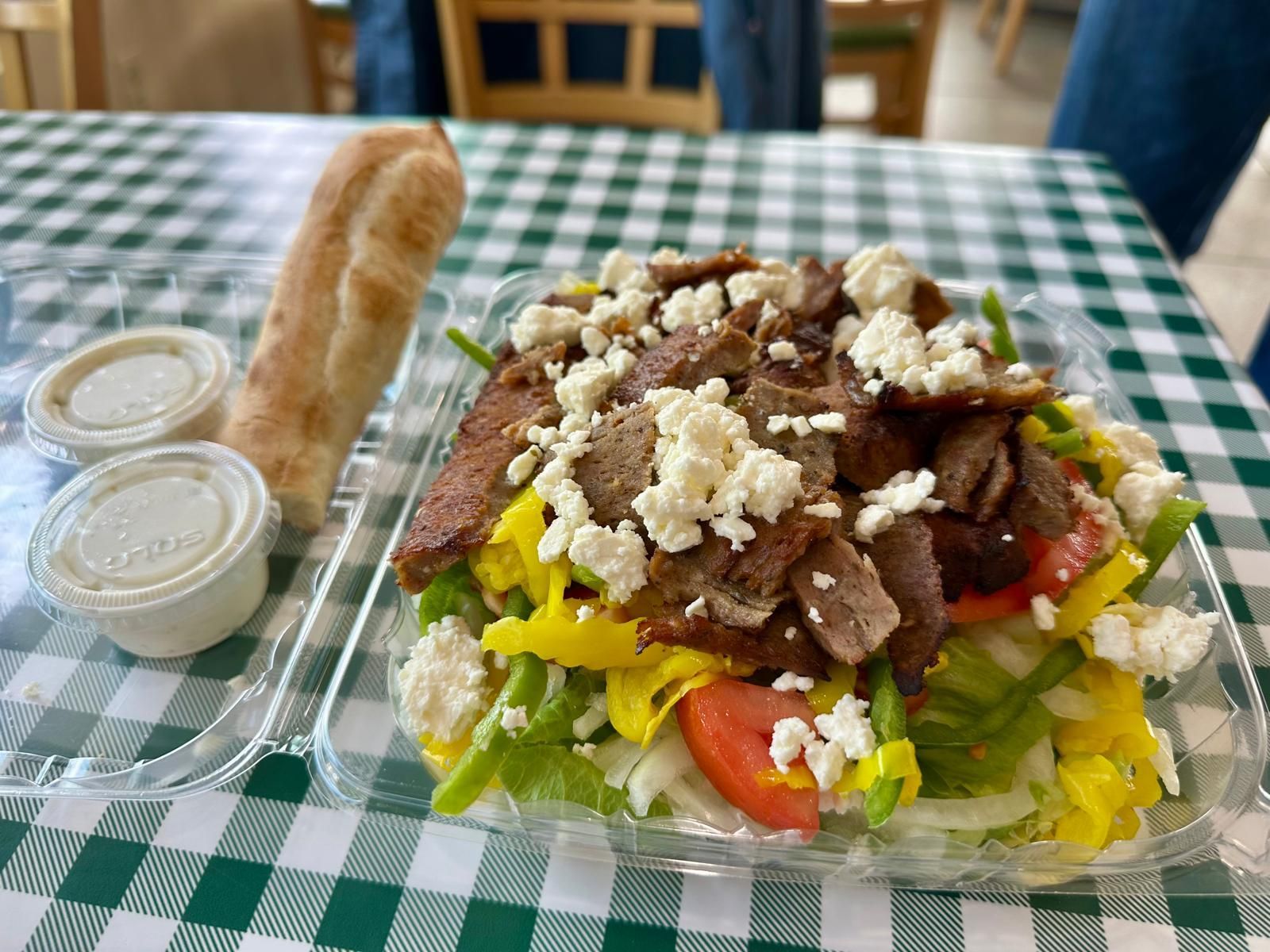 Salad with meat, feta cheese, tomatoes, and peppers, served with bread and dressing on a checkered table.
