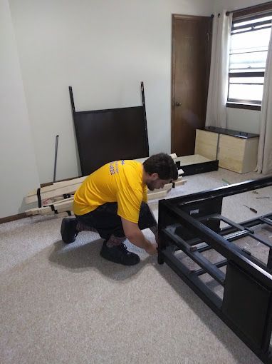 A man in a yellow shirt is kneeling down to install a bed frame in a bedroom.