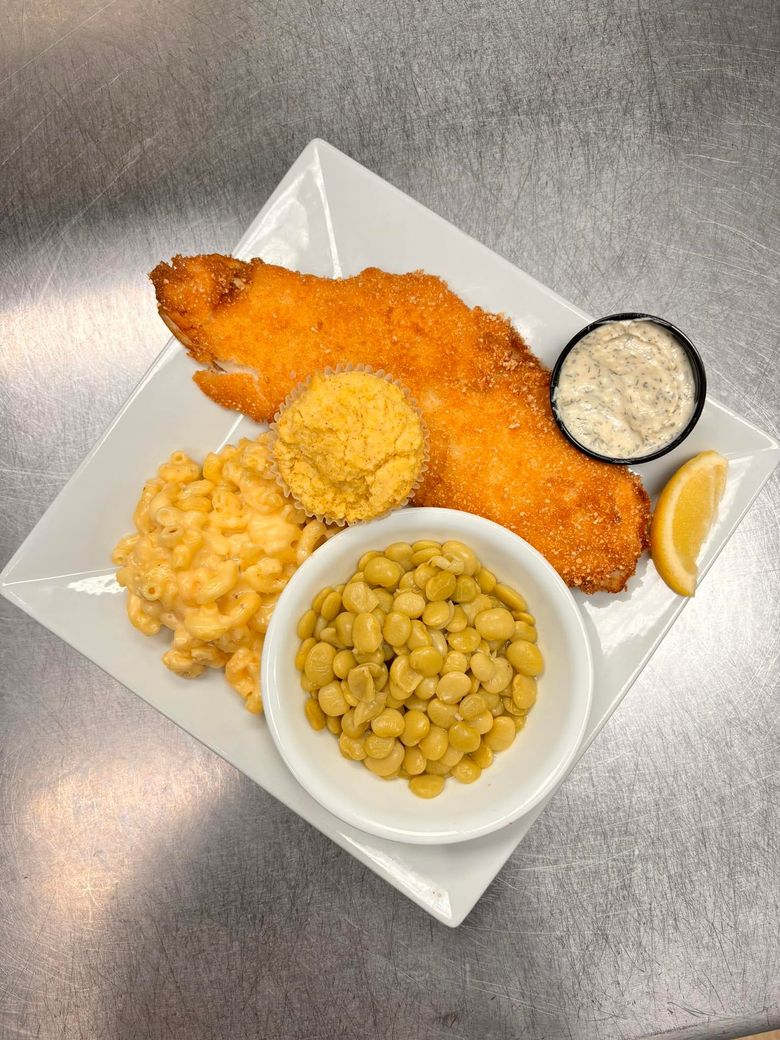 A breaded fish fillet served with mac and cheese, butter beans, a muffin, tartar sauce, and a lemon wedge on a white plate.