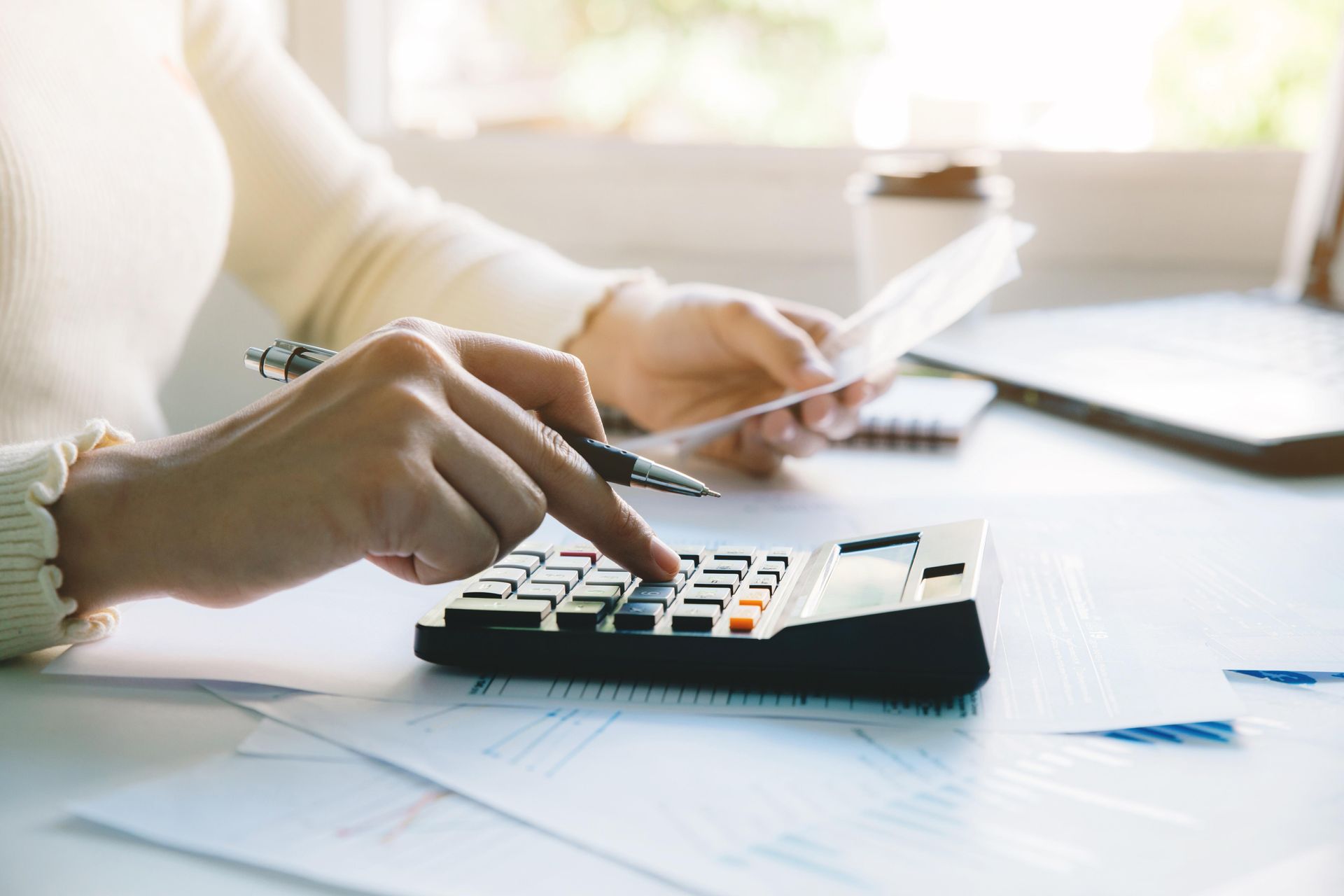 Person calculating expenses with a calculator, paperwork, and a coffee cup on a desk.