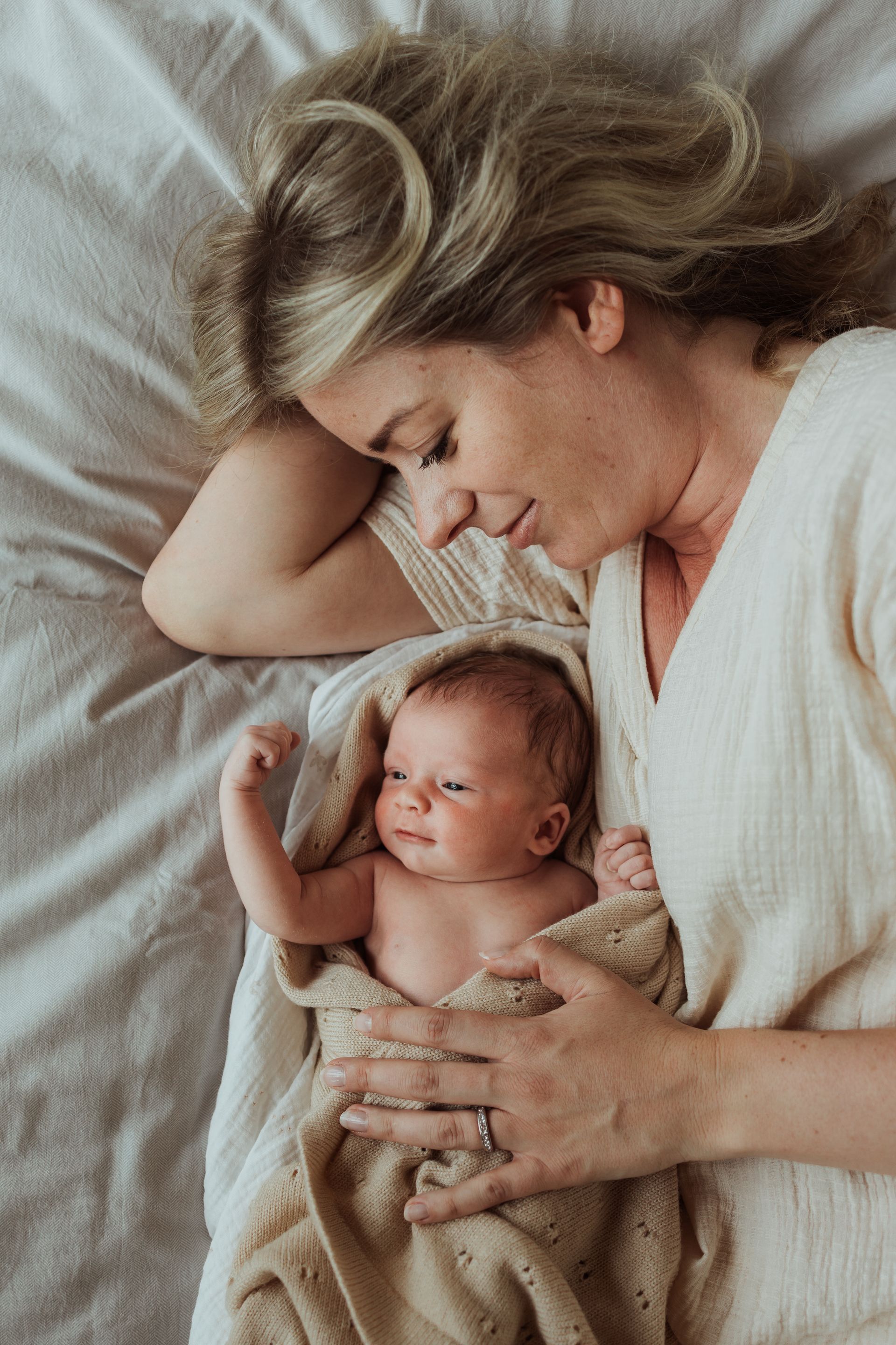 Een vrouw ligt op een bed met een pasgeboren baby in haar armen, gewikkeld in een deken.
