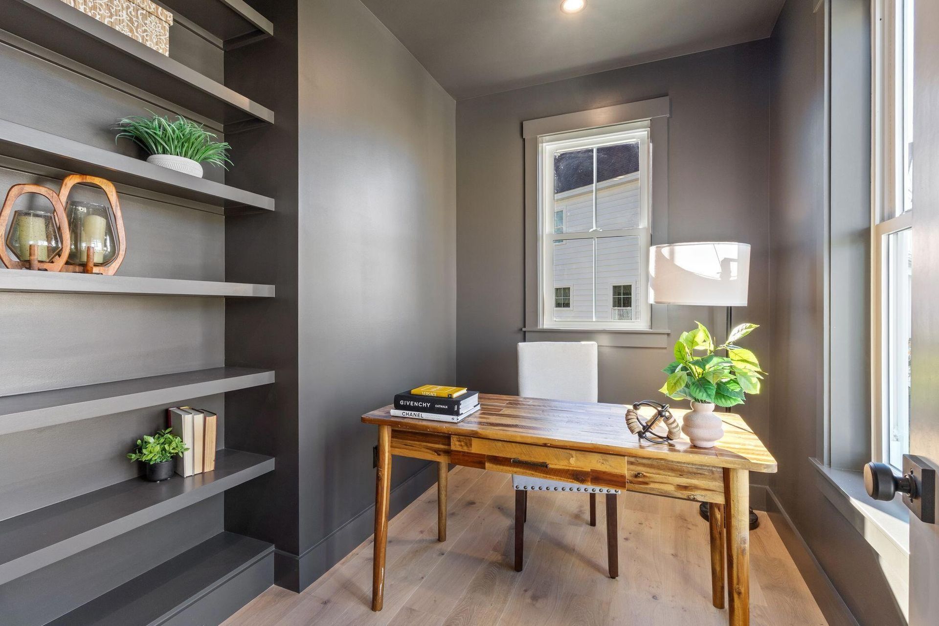 Modern home office with gray walls, wooden desk, shelves, and a window with plants and books.