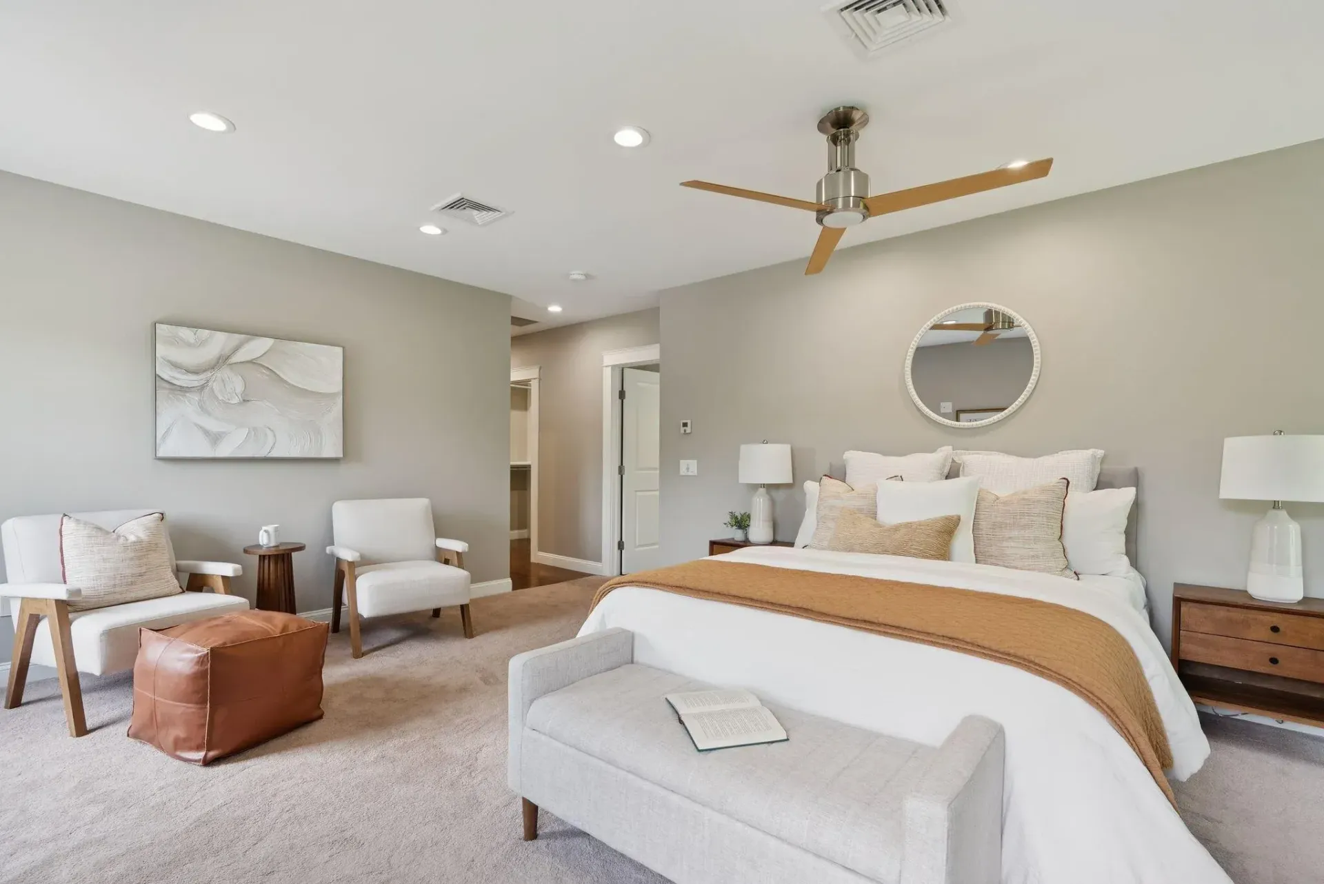 Bright bedroom with a bed, armchairs, ottoman, ceiling fan, and round mirror in neutral tones