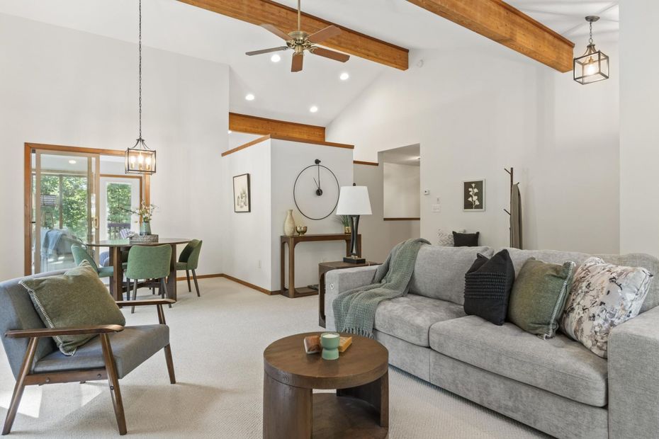 Bright living room with gray sofa, wood beams, green chairs, and a dining area by windows.