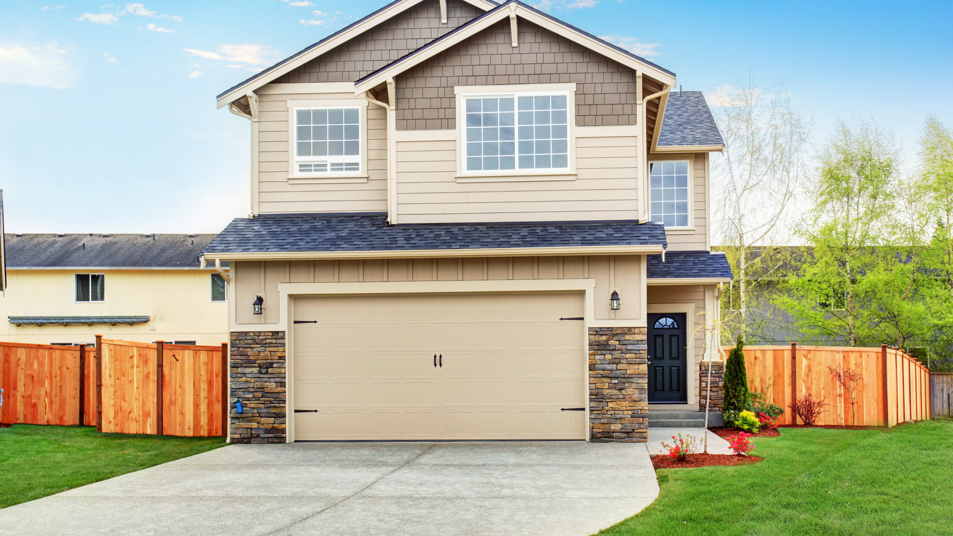 Two-story beige house with a tan garage door, set in front of a wooden fence on a sunny day.