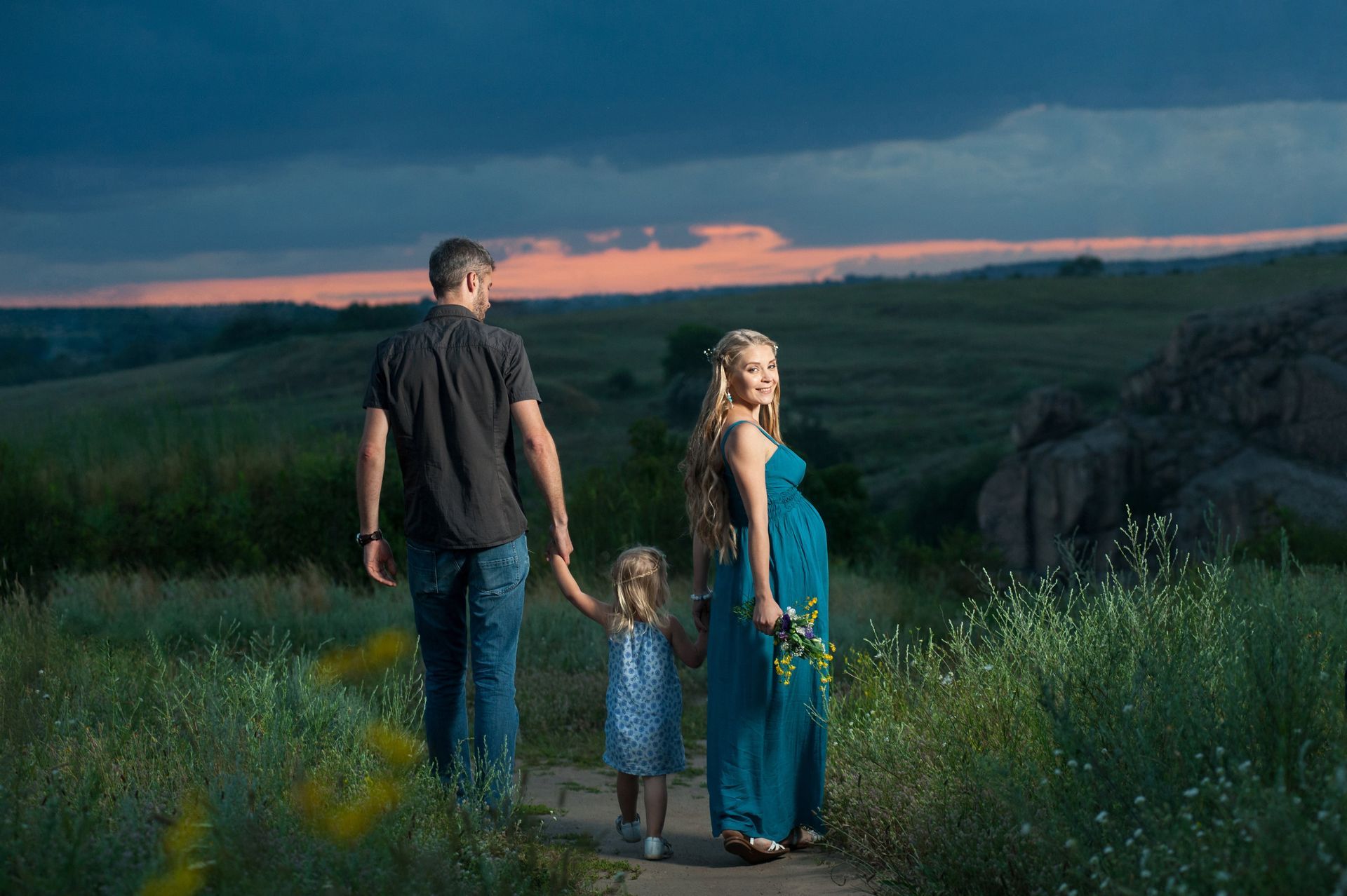 A pregnant woman , a man and a little girl are walking in a field.