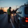 Roadside worker in reflective vest holding flashlight, next to a parked car at dusk.