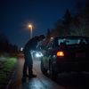 A person stands by a car at night, appearing to be looking at the door. Roadside setting with streetlights.