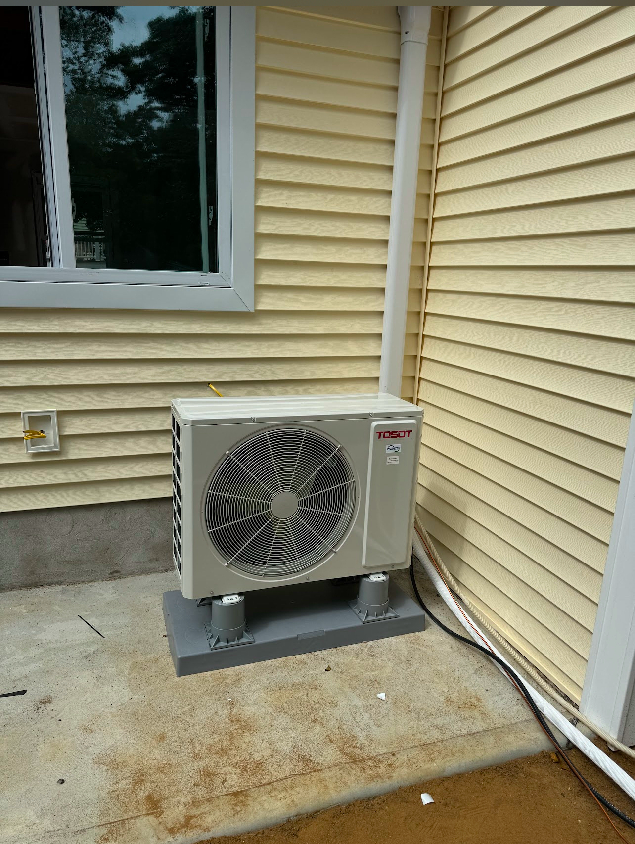 A white air conditioner is sitting on the side of a house next to a window.