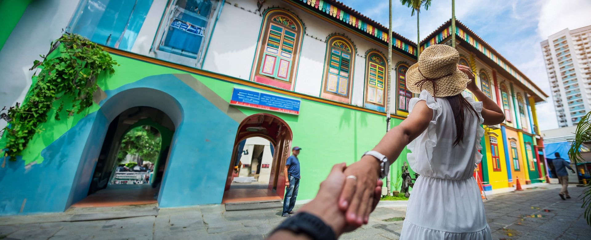 Una mujer con un vestido blanco sostiene la mano de un hombre frente a un edificio colorido.
