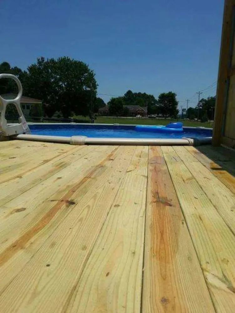 Wooden deck beside an above-ground pool under a clear blue sky, with trees in the background