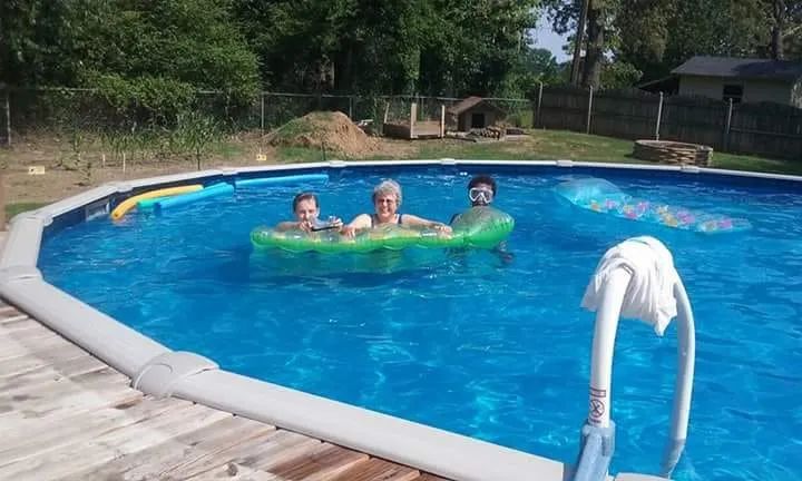 Three people relax on a green float in a blue backyard pool near a wooden deck.