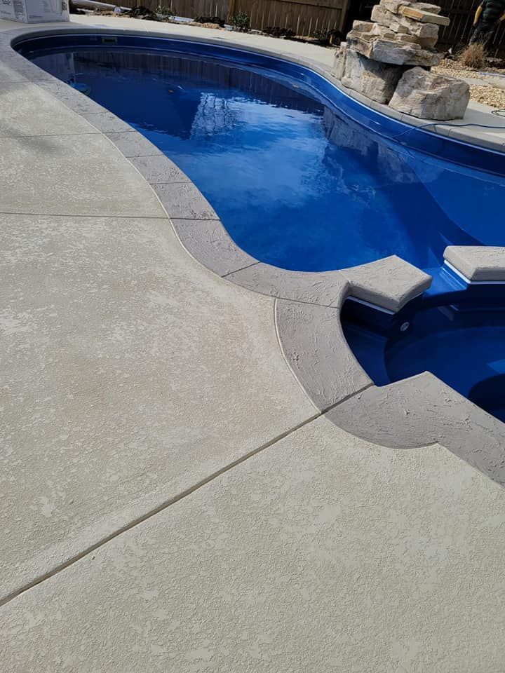 Curved concrete pool deck beside a blue in-ground swimming pool with stone waterfall feature