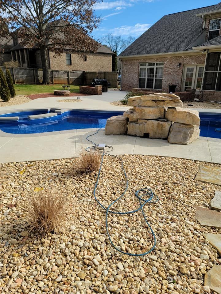Backyard pool with stone water feature, patio, hose, and rock landscaping under sunny skies