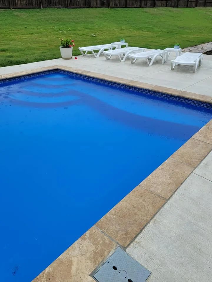 Blue swimming pool beside white lounge chairs on a patio with a grassy lawn.