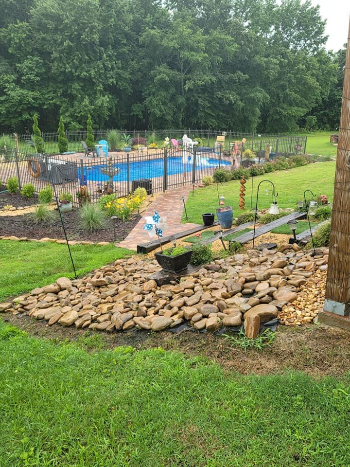 Backyard with a fenced blue pool, garden beds, stone walkway, and rock-lined slope under cloudy skies