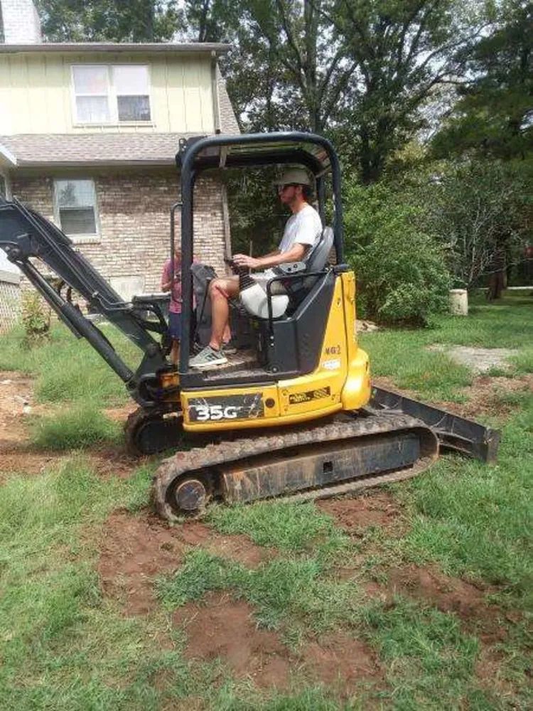 Yellow mini excavator with an operator working in a yard near a house and trees.