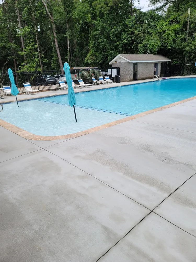 Outdoor swimming pool with blue umbrellas, lounge chairs, and a small shed beside a wooded area