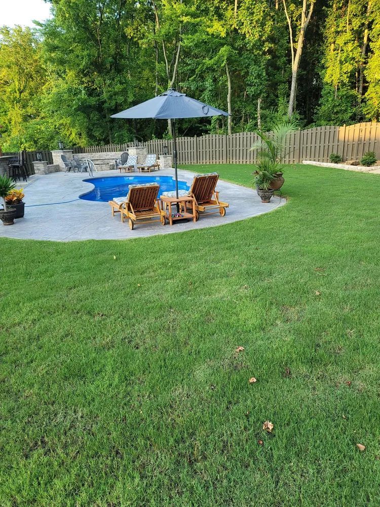 Backyard patio with blue umbrella, wooden chairs, and a round pool surrounded by green lawn and trees