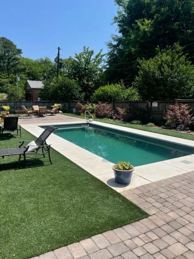 Backyard pool with lounge chairs, potted plant, and green lawn beside a brick patio.